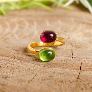 May include: A gold-colored ring with two oval gemstones. One gemstone is a deep red color, and the other is a vibrant green. The ring's design features a swirling band that holds the gemstones in place, creating a unique and elegant look.