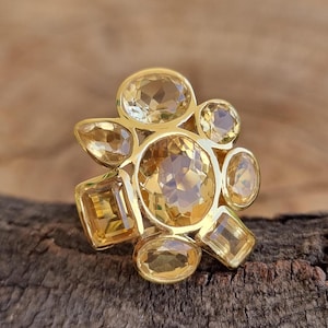 Può includere: Un anello dorato con diverse pietre preziose gialle di varie forme, tra cui ovali, rotonde e rettangolari. Le pietre sono disposte in un design a grappolo, creando un aspetto floreale. L'anello è esposto su una superficie di legno.
