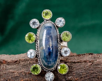 May include: A silver ring featuring a large, oval blue gemstone at the center, surrounded by smaller, round gemstones in shades of green and light blue. The ring is set on a textured wooden surface.