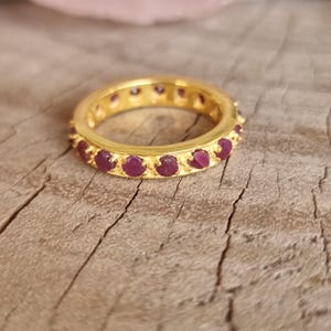 Könnte beinhalten: Ein goldfarbener Bandring mit einer durchgehenden Reihe kleiner, runder, tief rosa-roter Edelsteine, die in einzelnen Krappen gefasst sind. Der Ring hat eine polierte metallische Oberfläche und eine gleichmäßige, kreisförmige Form.