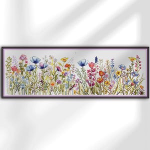 Op de afbeelding: Een ingelijste kruissteek van een wilde bloemenweide. Het kunstwerk toont een verscheidenheid aan kleurrijke bloemen in tinten blauw, roze, geel en oranje, tegen een witte achtergrond. De lijst is donker.