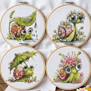 May include: Four cross-stitch embroidery hoops with charming snail designs. Each snail has a colourful shell and is decorated with flowers and leaves. The snails are set against a white background, and the hoops are made of wood.
