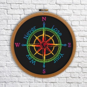 Puede incluir: Una brújula arcoíris con las palabras "Never Lose Your Way" alrededor de la brújula. La brújula está en un aro de bordado de madera.