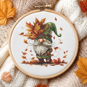 May include: Cross-stitch embroidery of a gnome wearing a green hat and holding autumn leaves. The gnome has a long white beard and is surrounded by falling leaves. The embroidery is set in a wooden hoop.