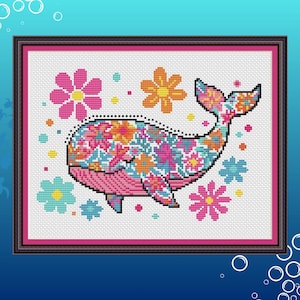 Peut inclure: Une broderie au point de croix encadrée représentant une baleine ornée de fleurs colorées. La baleine est rose et ornée de motifs floraux roses, oranges et bleus. Des fleurs roses et bleues entourent la baleine sur fond blanc.