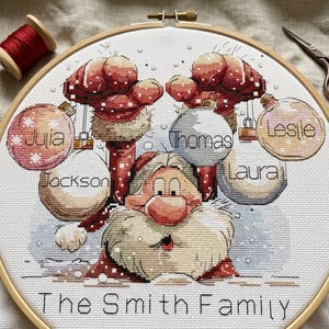 Puede incluir: Un bastidor de bordado de punto de cruz con un diseño de Papá Noel y el texto "The Smith Family". El diseño incluye adornos navideños con los nombres: Julia, Jackson, Thomas, Laura y Leslie. Una bobina de hilo rojo y unas tijeras están en el fondo.