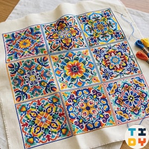 Puede incluir: Un trozo cuadrado de tela Aida de color blanco roto con un patrón de punto de cruz de nueve cuadrados distintos similares a azulejos. Cada cuadrado muestra un diseño floral y geométrico simétrico único en tonos vibrantes de azul, amarillo, naranja y rojo.