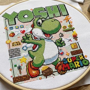 Può includere: Punto croce di Yoshi di Super Mario. Il dinosauro verde è in posa di corsa, con la scritta "YOSHI" sopra e "SUPER MARIO" sotto. Lo sfondo include elementi pixelati all'interno di un telaio da ricamo in legno.