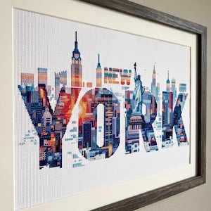 May include: A framed cross-stitch embroidery featuring the word NEW YORK in large block letters. The letters are filled with a colorful cityscape design, including the Statue of Liberty and skyscrapers in shades of blue, orange, and red.