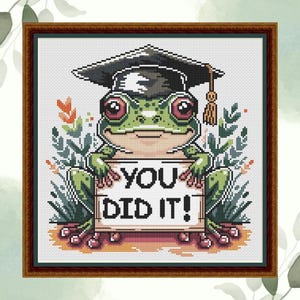 May include: Cross-stitch picture of a green frog in a graduation cap, holding a sign saying "YOU DID IT!". The frog is surrounded by plants, set in a brown frame. A celebratory piece of art.