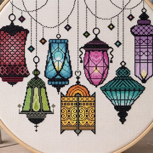 Può includere: Un ricamo a punto croce con sette lanterne colorate di varie forme e motivi. Le lanterne sono in tonalità di rosso, blu, verde, giallo, rosa e viola, sospese da una linea tratteggiata con accenti a forma di diamante. Il ricamo è incorniciato in un cerchio di legno.