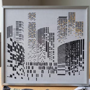 May include: A framed cross-stitch artwork depicting a stylized city skyline. The design features black and grey thread forming geometric skyscraper silhouettes against a white fabric background, with small yellow accents representing lit windows.