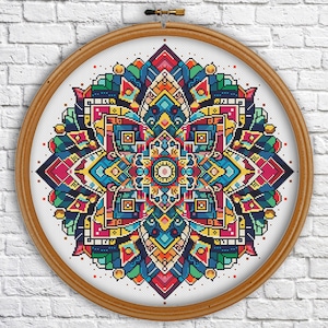Pode incluir: Um design de mandala colorido em ponto cruz num bastidor de bordar de madeira. A mandala apresenta um padrão simétrico com formas geométricas em tons de vermelho, azul, amarelo, verde e laranja. O bastidor é exibido contra uma parede de tijolos brancos.