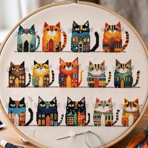 May include: A cross-stitch embroidery hoop with a pattern of colourful, house-like cats. The cats are in various colours, including blue, orange, and green, and are arranged in rows on a cream-coloured fabric. The hoop is made of wood.