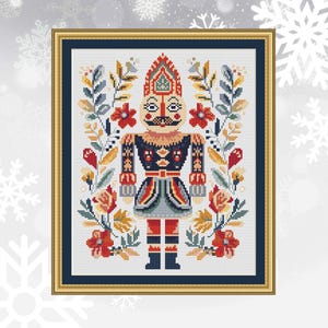May include: A cross-stitch pattern featuring a colorful nutcracker with a red, blue, and yellow hat, surrounded by floral designs in shades of red, yellow, and green.