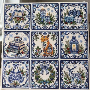 May include: A square cross-stitch sampler featuring nine distinct panels with blue and white motifs, including birds, candles, pumpkins, books, a fox, a lantern, mittens, a wreath, and a deer, all framed by intricate blue floral borders.