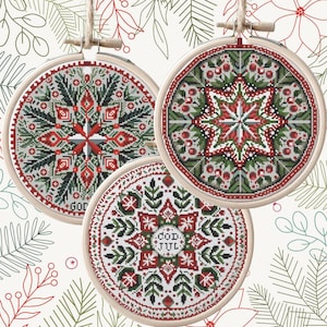 May include: Three round cross-stitch decorations in wooden hoops. Each has a red, green, and white geometric pattern. The ornaments are suspended by twine against a backdrop of Christmas-themed foliage.