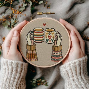 Puede incluir: Patrón de punto de cruz de dos manos sosteniendo tazas de café o té. Las tazas están decoradas con un diseño festivo de invierno. Las manos llevan guantes acogedores y con patrones.