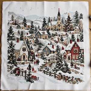May include: A cross-stitch embroidery on white Aida fabric depicting a snowy winter village scene. The design features multiple houses, a church with a steeple, evergreen trees, and small figures walking through the snow-covered landscape.