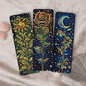 Puede incluir: Tres marcapáginas rectangulares con diseños pixelados. Uno presenta un sol, otro una rosa y el tercero una luna creciente. Cada uno tiene un tallo con hojas sobre un fondo azul oscuro estrellado. El sol y la luna son amarillos, y la rosa es roja.