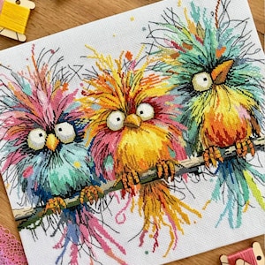 May include: A cross-stitch embroidery piece on white fabric featuring three colorful, fluffy birds with large eyes perched on a branch. The birds are rendered in vibrant shades of pink, blue, yellow, and orange with messy, textured plumage.