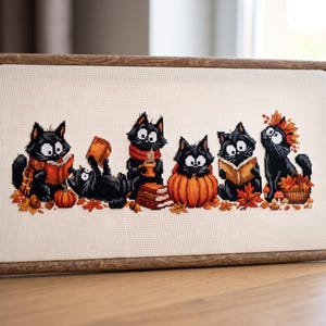 Könnte beinhalten: Ein rechteckiges Kreuzstich-Stickbild in einem Holzrahmen mit sechs schwarzen Katzen mit großen weißen Augen. Die Katzen sind in einer herbstlichen Umgebung mit orangefarbenen Kürbissen, braunen Büchern und herbstlichen Blättern dargestellt.