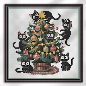 Op de afbeelding: Kruissteekkunst met een kerstboom versierd met ballen en lichtjes, omringd door vijf zwarte katten. De boom staat in een geweven mand op een patroonkleed, omlijst door een donkere houten rand.