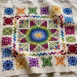 Könnte beinhalten: Ein quadratisches Stück beigefarbener Leinenstoff mit einem großen, mehrfarbigen Kreuzstich-Sternmuster in der Mitte, umgeben von einer Zickzack-Borte und kleineren, einzelnen geometrischen Sternmotiven in verschiedenen Farben wie Blau, Grün und Rot.