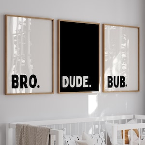 May include: A nursery scene featuring a white crib with bedding, pillows, and a decorative banner. Above the crib, three framed prints display the words "BRO.", "DUDE.", and "BUB." in bold black letters. A woven basket and a lamp are also visible.