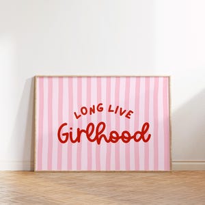 Op de afbeelding: Een ingelijste print met een roze en witte gestreepte achtergrond. De woorden "LONG LIVE Girlhood" zijn in rood script geschreven. De lijst is licht van kleur en de print wordt op een witte muur getoond.