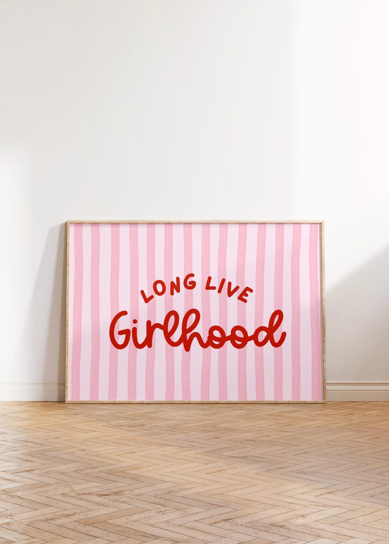 Op de afbeelding: Een ingelijste print met een roze en witte gestreepte achtergrond. De woorden "LONG LIVE Girlhood" zijn in rood script geschreven. De lijst is licht van kleur en de print wordt op een witte muur getoond.
