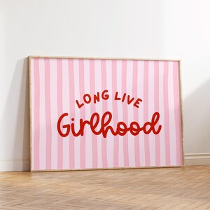 Op de afbeelding: Een ingelijste kunstafdruk met een roze en witte gestreepte achtergrond. De woorden "LONG LIVE Girlhood" zijn in rode script geschreven. Het kunstwerk wordt op een houten vloer getoond.
