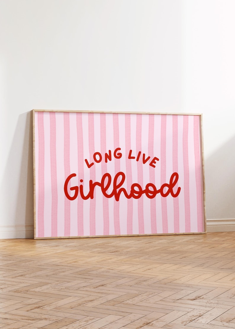Op de afbeelding: Een ingelijste kunstafdruk met een roze en witte gestreepte achtergrond. De woorden "LONG LIVE Girlhood" zijn in rode script geschreven. Het kunstwerk wordt op een houten vloer getoond.