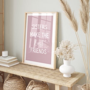 May include: A framed print with a pink background and white text that reads "Sisters make the best friends". The print is sitting on a light brown wicker bench with a white vase and dried flowers.