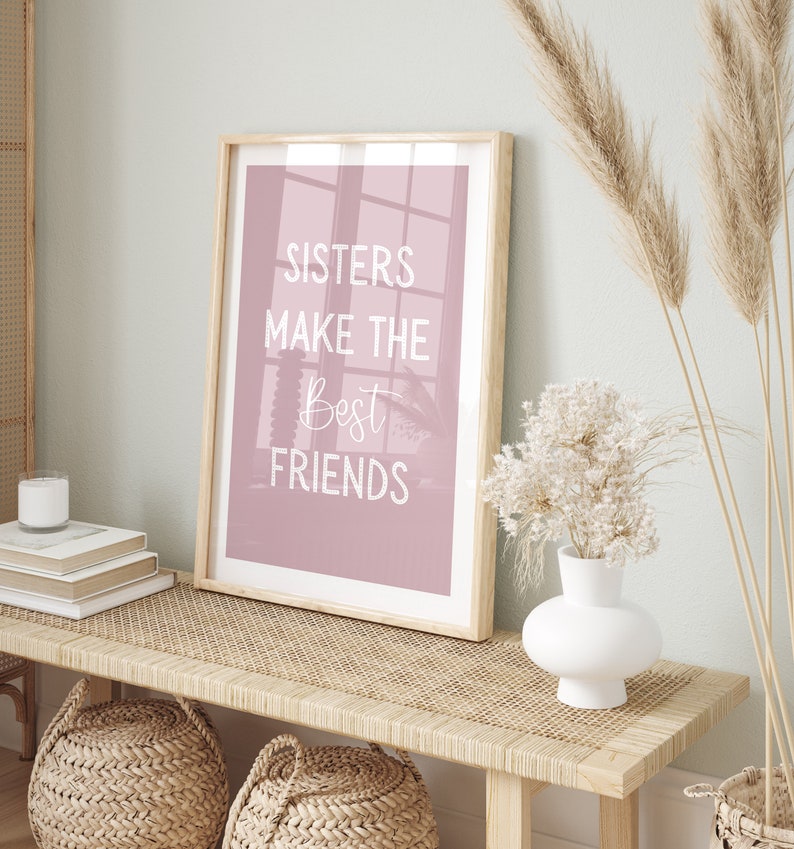 May include: A framed print with a pink background and white text that reads "Sisters make the best friends". The print is sitting on a light brown wicker bench with a white vase and dried flowers.