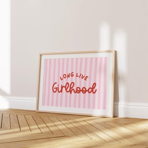 Op de afbeelding: Een ingelijste print met de woorden "Long Live Girlhood" in rood script. De achtergrond heeft verticale roze en witte strepen. De lijst is licht van kleur en de print leunt tegen een witte muur op een houten vloer.
