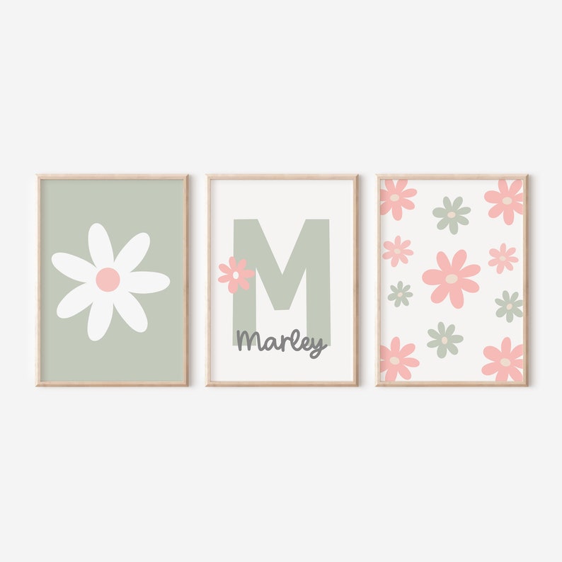 May include: Three framed prints with a green background. The first print features a white daisy with a pink center. The second print features a large green letter M with a small pink daisy in the corner and the name "Marley" written below. The third print features a pattern of pink and green daisies on a white background.