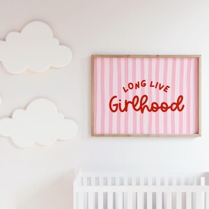 Op de afbeelding: Een ingelijste print met de woorden "LONG LIVE Girlhood" in rode letters op een roze gestreepte achtergrond. Witte wolk-vormige wanddecoratie en een wit ledikant zijn ook zichtbaar.