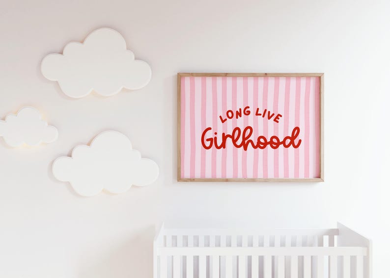 Op de afbeelding: Een ingelijste print met de woorden "LONG LIVE Girlhood" in rode letters op een roze gestreepte achtergrond. Witte wolk-vormige wanddecoratie en een wit ledikant zijn ook zichtbaar.