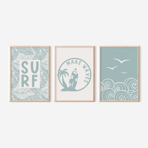 May include: Set of three blue and white framed prints with a beach theme. The first print features a palm tree and surfboard with the text "MAKE WAVES". The second print features a leafy pattern with the text "SURF". The third print features a blue ocean with white waves and white seagulls.