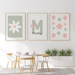 May include: Three framed prints with a green background and pink flowers. The center print features the letter "M" and the name "Marley". The prints are hanging on a white wall above a white table and chairs.