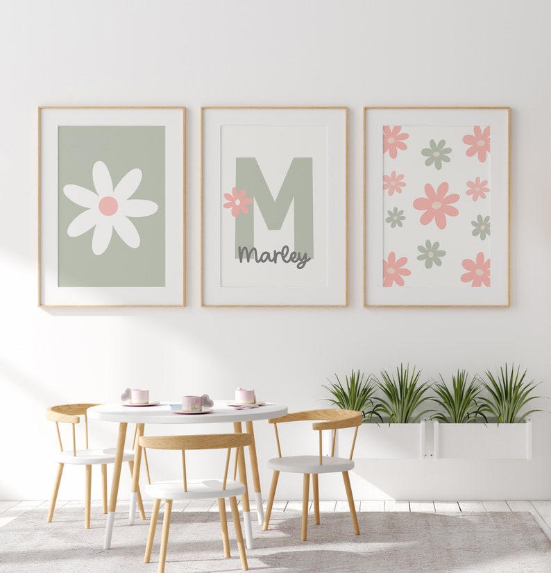 May include: Three framed prints with a green background and pink flowers. The center print features the letter "M" and the name "Marley". The prints are hanging on a white wall above a white table and chairs.