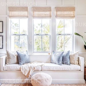 Puede incluir: Un asiento de ventana blanco con un cojín blanco y almohadas azules. Una manta blanca está drapeada sobre el cojín. Un puf blanco se encuentra frente al asiento. Una gran planta verde en una cesta tejida está en la esquina de la habitación.