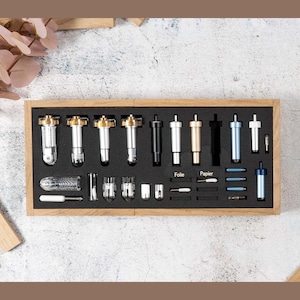 Może przedstawiać: Drewniane pudełko zawierające różne ostrza tnące do maszyny do cięcia rękodzieła. Ostrza są uporządkowane według typu i koloru, z etykietami „Folie” i „Papier”.