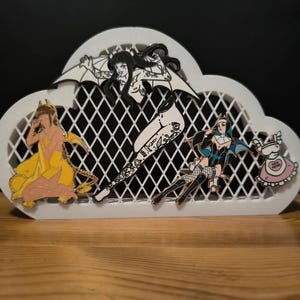 Puede incluir: Porta-pins en forma de nube blanca con un pin blanco y negro de una mujer con alas, un pin amarillo de una mujer con una corona, un pin azul de una mujer con alas y un pin rosa de una mujer con un corazón.