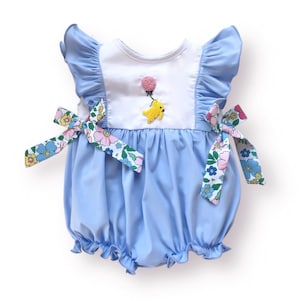 May include: A light blue baby romper with white accents and floral print bows. The romper features a ruffled neckline and sleeves, and an embroidered yellow chick holding a pink balloon. The romper is designed for infants and toddlers.