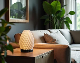 May include: A white ceramic lamp with a swirling design sits on a wooden table in a living room. The lamp is turned on and emits a warm glow. The lamp is in front of a beige sofa with a throw pillow.