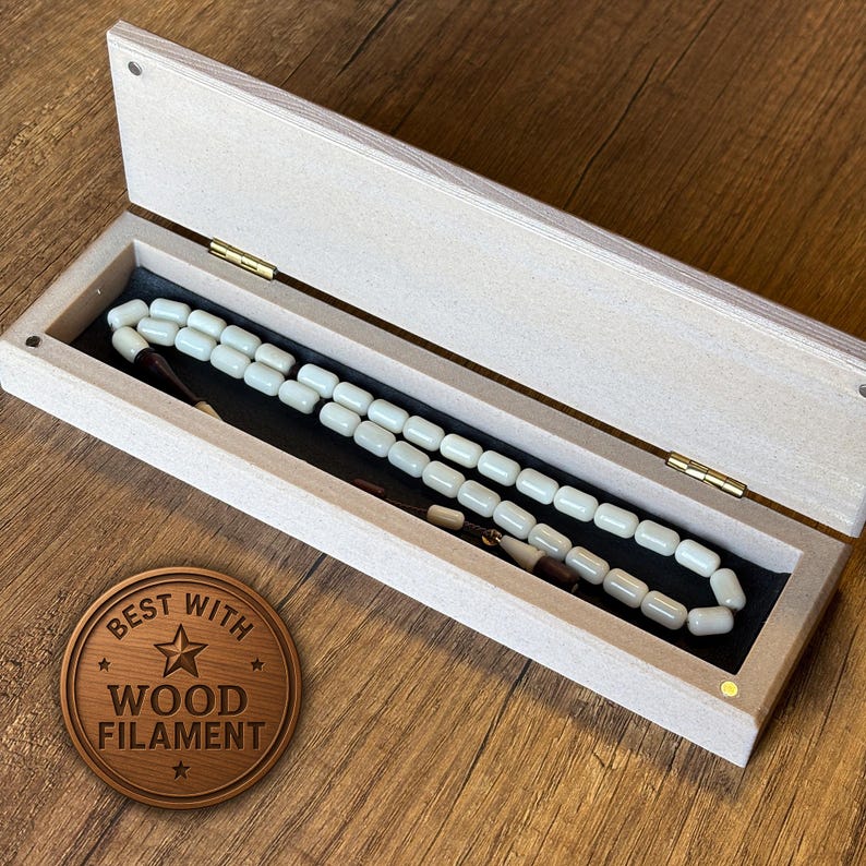 Peut inclure: Une bo&icirc;te en bois de couleur claire avec un couvercle &agrave; charni&egrave;re, contenant un chapelet de perles blanc cass&eacute;. Les perles sont rectangulaires et enfil&eacute;es ensemble. La bo&icirc;te est doubl&eacute;e de tissu noir. Un m&eacute;daillon en bois avec le texte "BEST WITH WOOD FILAMENT" est visible.