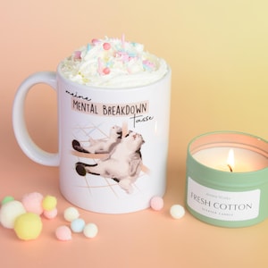 Könnte beinhalten: Weiße Keramiktasse mit einer Katzenillustration und dem Text "meine MENTAL BREAKDOWN Tasse". Die Tasse ist mit Schlagsahne und bunten Streuseln gefüllt. Im Hintergrund befindet sich eine grüne Kerze mit dem Text "Fresh Cotton".