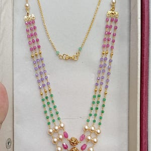 May include: A multi-strand necklace featuring three layers of beads in pink, purple, and green, accented with cream-colored pearls and three central gold-toned filigree spheres. The necklace is connected by a thin gold-toned chain with a lobster clasp.